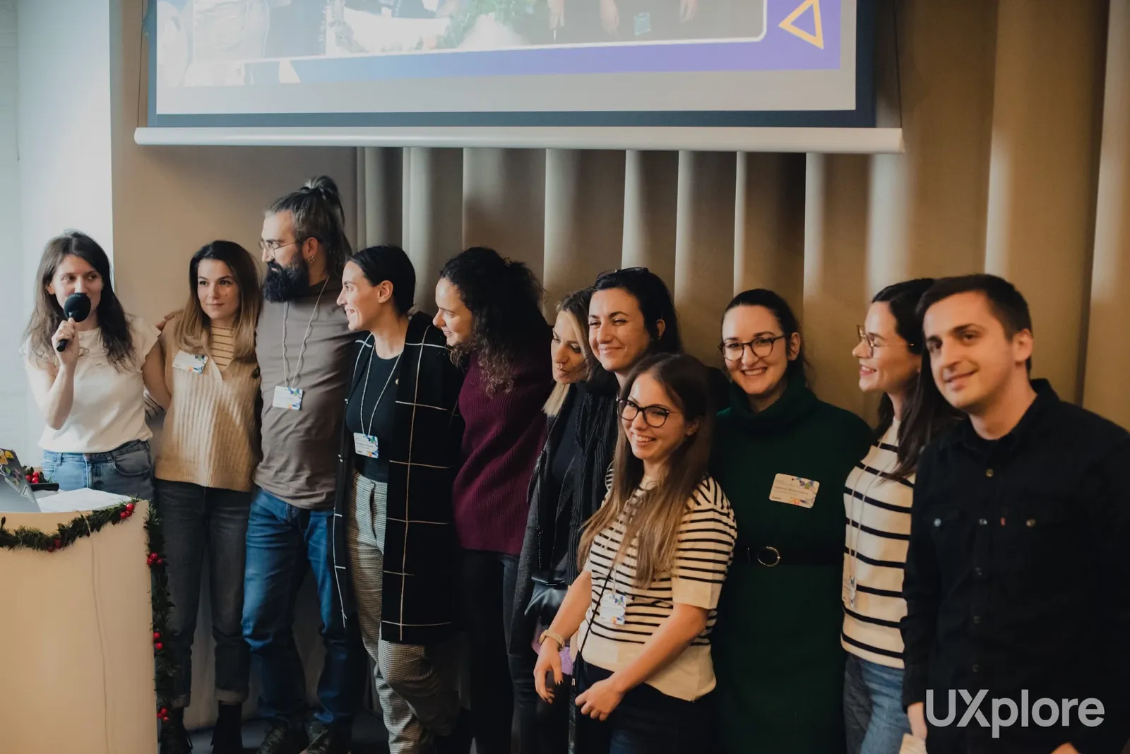 Ten members of the UXplore team standing close together in a row, smiling, in front of a curtain wall, one of them holding a microphone, with a projected slide above them.